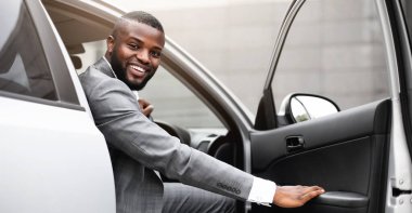 Pahalı takım elbiseli neşeli siyah adam lüks arabaya biniyor, kapıyı tutuyor ve kameraya gülümsüyor. Afrikalı Amerikalı başarılı girişimci sabah ofise gidiyor.