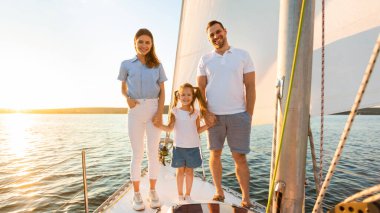 Deniz kenarında yaz tatili. Tekne güvertesinde duran gülümseyen aile hafta sonunun tadını çıkartıyor. Veliler ve kızı el ele tutuşup yelkenlideki kamera pozlarına bakıyorlar.