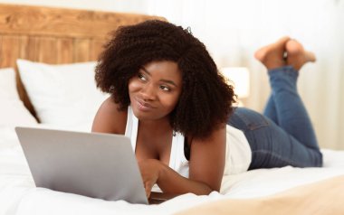 Evde dizüstü bilgisayarıyla çalışan güzel siyah kız öğrenci yatakta uzanıp düşünceli bir şekilde havaya bakıyor.