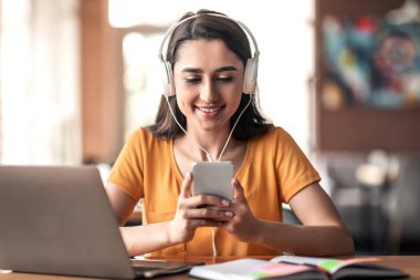 Kulaklık takmış genç bir kız, kafede oturuyor, dizüstü bilgisayar ve akıllı telefon kullanıyor.