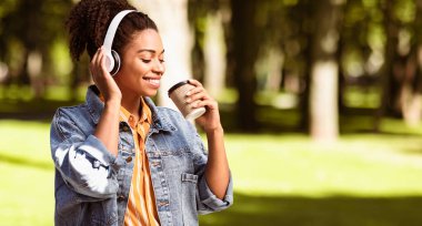 Hafta sonu konsepti. Kulaklıklı Afrikalı Milenyum Kızı dışarıda parkta kahve içip eğleniyor. Panorama, Boşluğu Kopyala