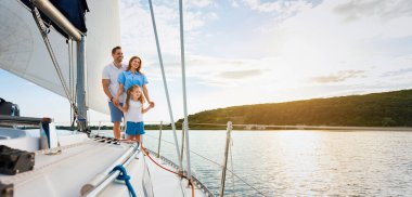 Neşeli bir aile güvertede durmuş güzel bir yaz günü deniz kenarında tatil yaparak yat gezintisinin tadını çıkarıyor. Ebeveynler ve Kız Hafta Sonu Yelkenliyle Açık Hava Gezisi. Panorama, Boş Alan