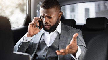 Kızgın Afrikalı Amerikalı iş adamı cep telefonuyla konuşuyor ve iş toplantısına arabayla giderken laptop ekranına bakıyor, genç siyah yönetici arka koltukta oturuyor ve çalışıyor, yakın plan çekiyor.