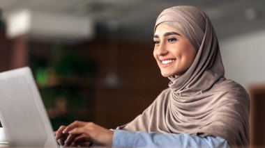 Kafe 'de çalışan tesettürlü neşeli Arap kadın dizüstü bilgisayar kullanıyor, kopyalama alanı kullanıyor. Gülümseyen Müslüman kadının bilgisayarında daktilo yazması, yeni uzak işinin keyfini çıkarması, müşterilerle sohbet etmesi.