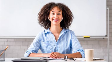 Kullanılabilir İngilizce dersleri var. Kablosuz kulaklıklı, gülümseyen Afro-Amerikan kadın iş yerinde oturup öğrencilere video çekiyor.