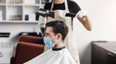 Koruyucu maskeli genç adam berber dükkanında saçını kestiriyor.