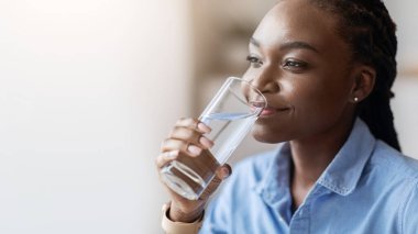 Sıvı kaybı. Güzel Afrikalı Amerikalı kadın bardaktan maden suyu içiyor ve uzaklara bakıyor, sağlıklı yaşam tarzının tadını çıkarıyor, seçici odaklanma ile yakın çekim yapıyor ve uzayı kopyalıyor.