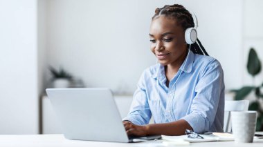 Milenyum Siyah Kadın Çalışanı Laptop ile çalışıyor ve ofiste müzik dinliyor, kulaklık takıyor, Masasında oturan Afrikalı kadın Bilgisayar klavyesinde oturuyor, e-postaları cevaplıyor, Boşluğu kopyalıyor