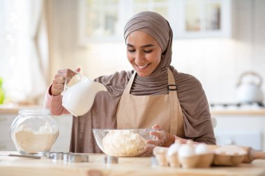 Ev Fırını. Mutfakta turta için hamur hazırlayan, kâseye malzeme ekleyen, önlük takan, ev yapımı yemek yapmaktan zevk alan, ev yapımı yemek yapmaktan hoşlanan, mutlu, genç Müslüman kadın.