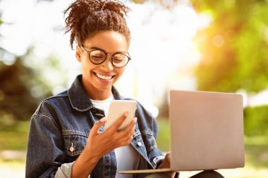 Akıllı telefondan mesaj atan Afrikalı Öğrenci Kız dışarıda parkta dizüstü bilgisayar kullanarak öğreniyor. E-Öğrenme Konsepti