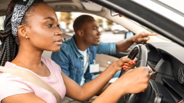 Konsantre genç siyahi kadın sürücü kursunda park pratiği yapıyor, yakışıklı bir eğitmenle arabada oturuyor. Milenyum Afrikalı Amerikalı erkek, kadın öğrencilere araba sürmede yardım ediyor.