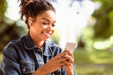 Sosyal Medya Sohbet Uygulamasında Akıllı Telefon Mesajı Kullanan Mutlu Siyahi Kadın Dışarıda Park 'ta Oturuyor. Cep telefonu kavramı için çevrimiçi mesajlaşma ve cep telefonu uygulamaları