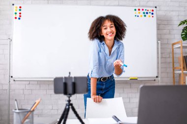 Uzaktan kumandalı İngiliz Webinar. Elinde keçeli kalemle mutlu bir Afrikalı Amerikalı kadın oturma odasının iç tarafındaki webcam 'e konuşuyor.