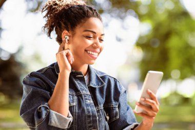 Afro-Amerikan kadın cep telefonu ve kulaklık kullanıyor. Dışarıdaki parkta video görüşmesi yapıyor. Modern Uzaktan İletişim, Cep Telefonu Kullanıcı Sohbeti Kulaklık Kullanıyor. Seçici Odaklanma