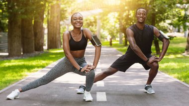 Siyah koşucu çift dışarıda kaslarını geriyor, parkta koşmadan önce ısınıyor, kameraya gülümsüyor, sağlıklı ve motive olmuş hissediyorlar, boş alan yaratıyorlar.