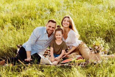Bağlanma kavramı. Neşeli aile dışarıda pikniğin tadını çıkarıyor, çimlerin üzerinde battaniyeye oturuyor ve kameraya bakıyor.