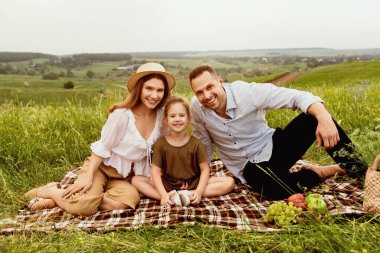 Tatil günü. Küçük çocukları battaniyeye oturmuş, arka planı bulanık bir şekilde kameraya bakan mutlu bir çift.