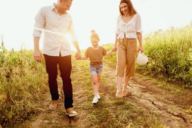 Bir çocukla zaman geçirmek. Mutlu bebek çayırda yürüyor ve gün batımında anne ve babasını elleriyle tutuyor.