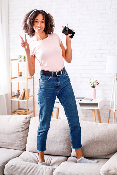 Любимая музыка дома во время самоизоляции. Счастливая африканская американка в наушниках танцует, поет и показывает знак мира на диване в интерьере гостиной