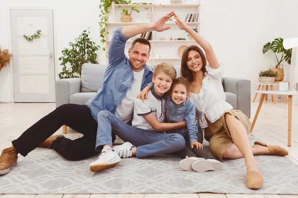 Aile Konutları. Mutlu Ebeveynler Evlerinde Zeminde oturan Çocuklarının Üzerinde Sembolik Çatı Yapıyor.