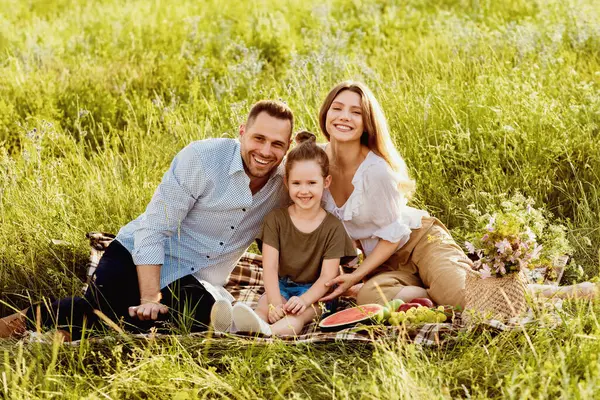Bağlanma kavramı. Neşeli aile dışarıda pikniğin tadını çıkarıyor, çimlerin üzerinde battaniyeye oturuyor ve kameraya bakıyor.