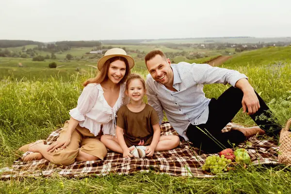 Tatil günü. Küçük çocukları battaniyeye oturmuş, arka planı bulanık bir şekilde kameraya bakan mutlu bir çift.