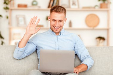 Neşeli adam video görüşmesi yapıyor, kucağında bilgisayarla oturuyor, el sallıyor, kulaklık takıyor.