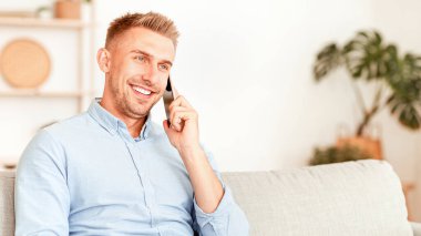 Mobil İletişim Konsepti. Neşeli bir adam telefon ediyor, ailesiyle ya da arkadaşıyla konuşuyor oturma odasında kanepede oturuyor. Kendine güvenen yetişkin çağrısı, kanepede dinlenme, boş alan