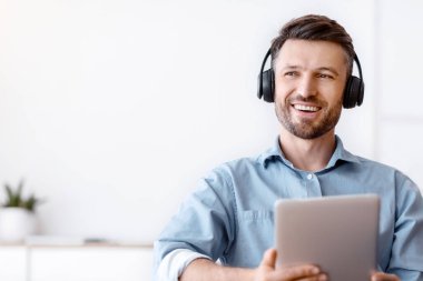 Yakışıklı, serbest çalışan, kablosuz kulaklıklı adam. Ofiste dijital tabletle rahatlıyor, işe ara veriyor, not defteriyle masada oturuyor, müzik dinliyor ve uzaklara bakıyor.