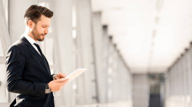 Yakışıklı sakallı iş adamı, uçuşu beklerken dijital tablet üzerindeki e-postaları kontrol ediyor, havaalanının penceresinin yanında duruyor, kopyalama alanı. Orta yaşlı girişimci şık takım elbiseli, ped kullanıyor.