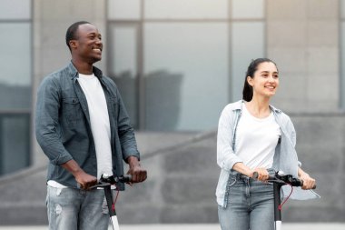 Canlı bir şehir ortamında iki genç dışarıda scooter kullanıyor. Gülümsüyor ve etkileşime giriyorlar, gençliğin neşesini ve çeşitli topluluklardaki açık hava aktivitelerini sergiliyorlar..