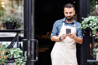 İnternette çiçekler hakkında blog, modern stüdyoda çiçekçilerin çalışmaları ve çiçek siparişi. Önlüklü neşeli genç sakallı Avrupalı adam farklı bitkilerle kırsal kesimlerin kapısında akıllı telefonuyla yazı yazıyor.