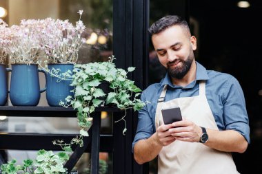 Çiçek merkezinin genç beyaz erkek sahibi portresi telefonda yazıyor, önlüklü çalışan müdürü envanter çıkarıyor. Neşeli adam saksılarda ve vazolarda bitkileri olan çiçekçinin yanındaki akıllı telefonu kullanıyor.