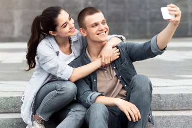 Mutlu sevgili konuşuyor. Merdivenlerde oturmuş selfie çekiyor. Ön tarafta iki tane morartılmış scooter var.