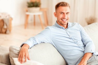 Dinlen ve rahatla kavramı. Gömlekli yakışıklı bir adamın portresi. Oturma odasında kanepeye yaslanıyor. Fotoğraf için poz veren tatmin olmuş adam, kameraya bakan, boş alan.