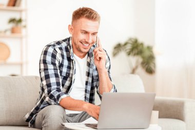 Yaşam tarzı kavramı. Başarılı menajerin portresi cep telefonuyla konuşuyor, bilgisayarda çalışıyor, modern dairede koltukta oturuyor. Evden işe gidiyorum. Çalışmak için dizüstü bilgisayar kullanan kendine güvenen bir adam, boş alan