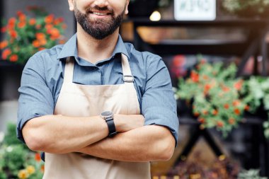 Stüdyo, kafe, dükkan sahibinin olumlu duyguları. Neşeli, kendine güvenen, önlüklü, göğsünde çapraz kolları olan sakallı yakışıklı bir adam. Çiçekçi arka planında, bitkilerle, kopyalama alanında.