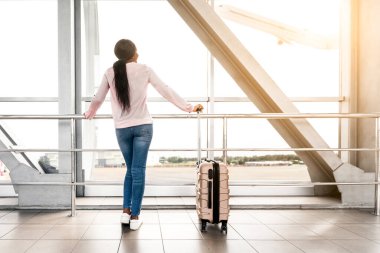 Air Transportation Concept. Happy Black Woman Looking At Plane While Waiting Flight In Airport, Young Traveller Lady Standing With Suitcase Luggage Near Window In Terminal, Enjoying Trip, Rear View