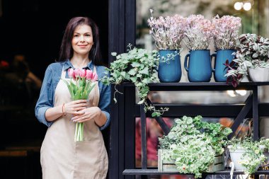 Startup, small business, eco restaurant outdoor and modern rustic flower shop. Smiling millennial beautiful female in apron holds bouquet of tulips for client, at front door of plants studio or cafe