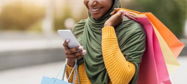 Taksi uygulaması. Gülümseyen Müslüman kadın, akıllı telefondan taksi çağırıyor, başarılı alışverişin ardından bir sürü parlak kağıt torba taşıyor, şoförü bekliyor, başörtüsü takıyor, özgür uzay giysisi giyiyor.