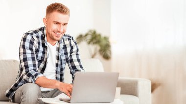 İçişleri Bakanlığı. Dizüstü bilgisayar kullanan, evdeki modern oturma odasındaki koltukta oturan, genç erkek alışverişinden memnun olan, çalışan, bilgisayar ekranına bakan, boş alan arayan sıradan bir adam.