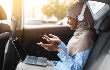 Çevrimiçi İletişim. Siyah Müslüman kadın, laptopuyla arabada video konferansı yapıyor, otomobilin arka koltuğunda oturuyor, bilgisayar ve kulaklıkla video görüşmesi yapıyor, cihaz kamerasında konuşuyor, yan görünüm