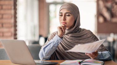 Düşünceli Müslüman kadın yönetici kafede otururken, laptop kullanıyor ve belgeleri kontrol ediyor, boşluğu kopyalıyor