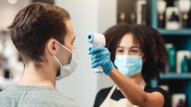 Salgın sırasında yeni iş kuralları kavramı. Kadın kuaför, berber dükkanındaki genç erkek müşterinin vücut ısısını ölçüyor.