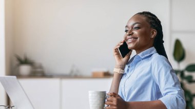 İşte rahatla. Modern ofisteki işyerinde cep telefonuyla konuşan ve kahve içen mutlu siyah sekreter.