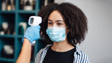 Koruyucu maskeli siyah kadın güzellik salonunda sıcaklık taraması yaptırıyor.