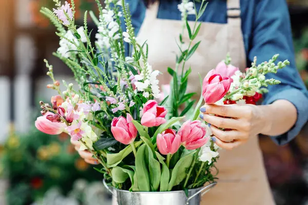 Doğum günü hediyesi, tarih, yıldönümü ve tatil, mağaza siparişi ve müşteri için çiçek tasarımı. Bin yıllık önlüklü kadın çiçekçi çiçek buketi yapıyor. Çiçekçi dükkanının yanındaki kovada lalelerle. Kırpılmış, açık havada.