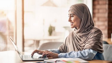Tesettürlü neşeli kız pencerenin yanındaki kafede oturuyor, internetten iş arıyor, dizüstü bilgisayar, yan görüş, boş alan kullanıyor.