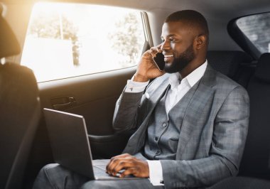 Mutlu siyah işadamı cep telefonuyla konuşuyor ve camdan bakıyor, dizüstü bilgisayar kullanıyor, iş toplantısına arabayla gidiyor, Afrikalı yönetici arabada oturuyor, telefonla konuşuyor.
