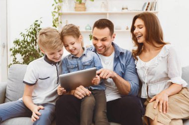 Mutlu ebeveynler ve çocuklar dijital tabletle oturup aile videolarının evde koltukta oturup gülüşünü izliyorlar. Hafta sonu Tatili